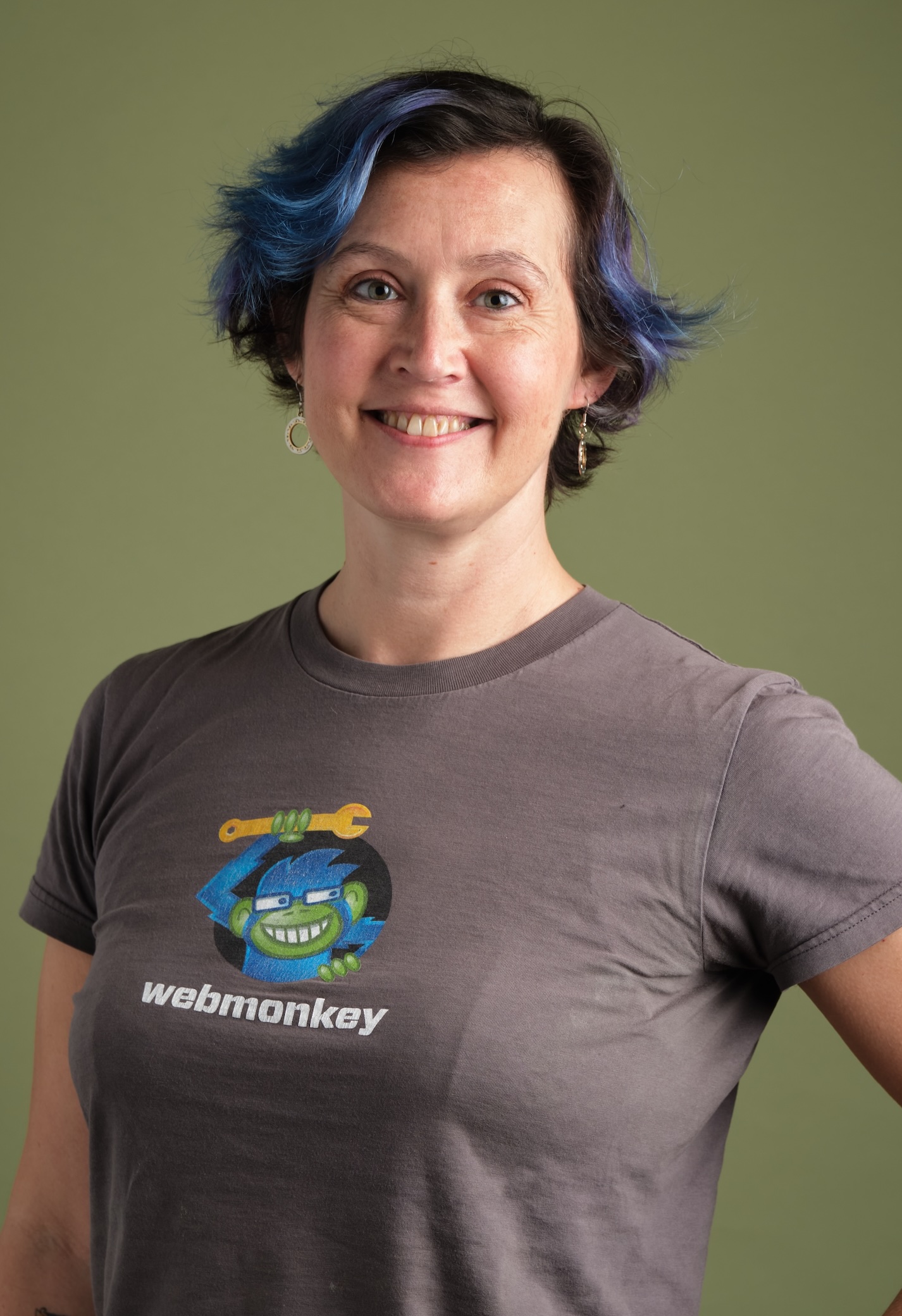 A headshot picture of Amy where her hair is short, brown and blue. She is smiling and has one hand on her hip.She is wearing a grey shirt that says Webmonkey and has their logo of a blue monkey with glasses holding an orange wrench. The background of the portait is an olive color.